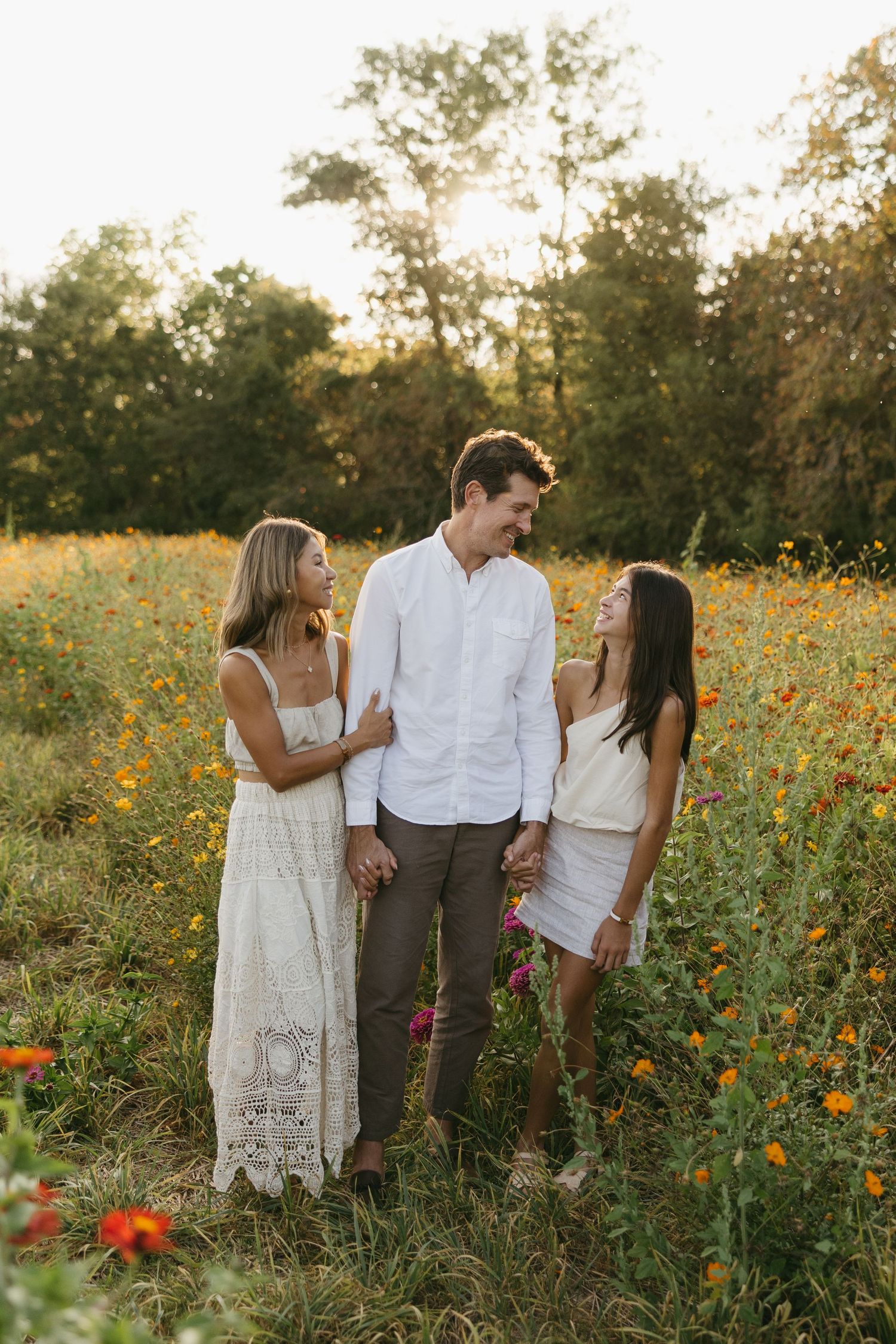 Smiling family of three standing together in a golden sunlit wildflower meadow.