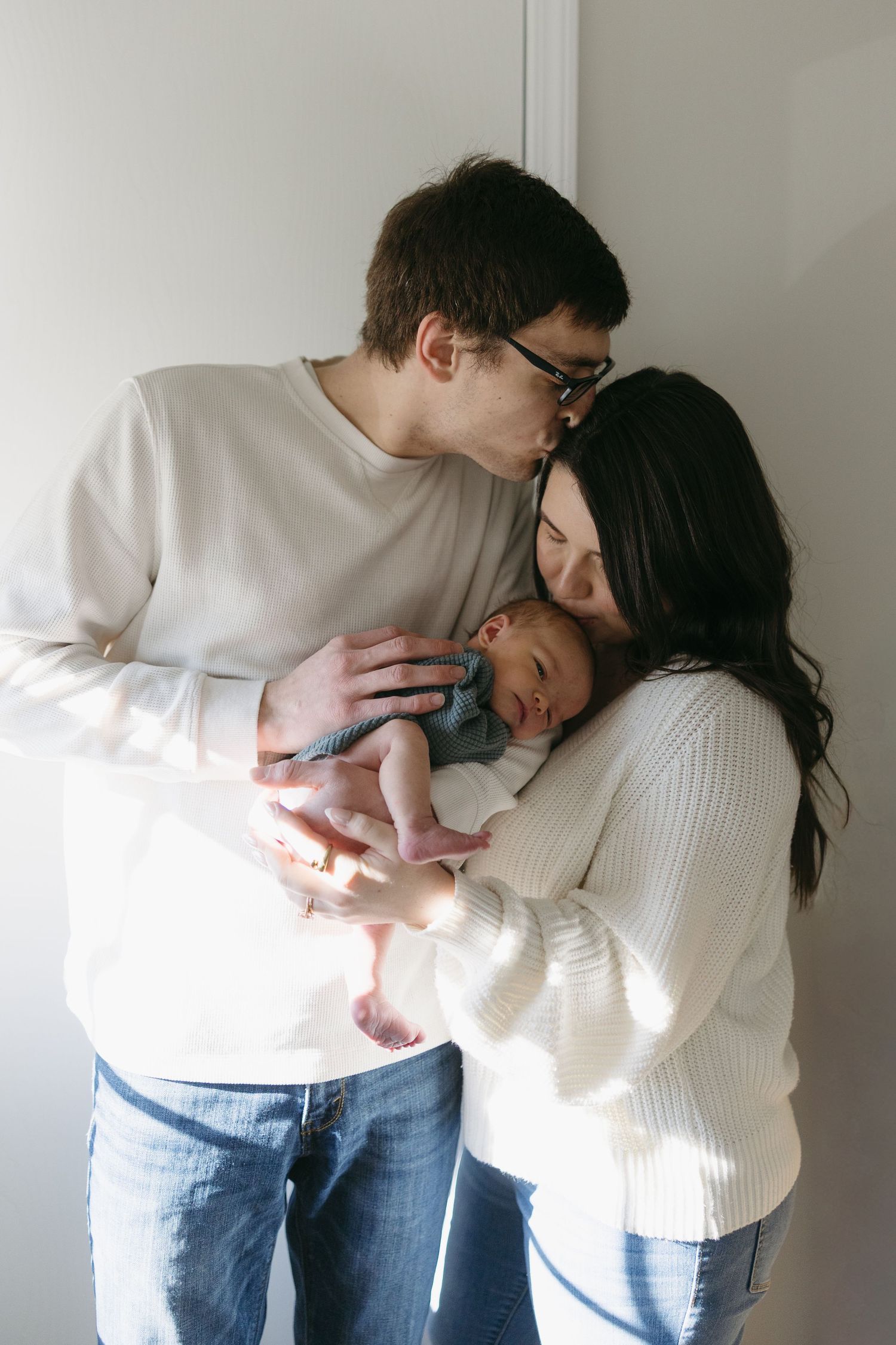 New parents tenderly kissing and cradling their sleeping newborn baby in soft natural light.