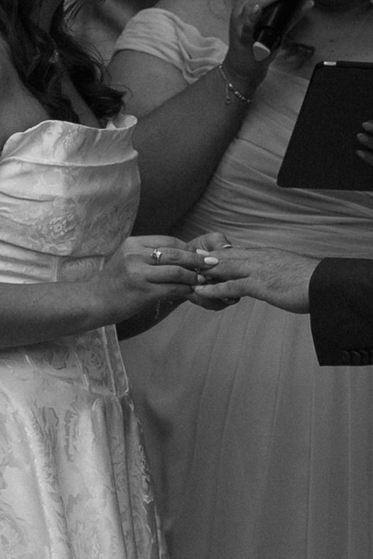 Black and white close-up of bride placing wedding ring on grooms hand during the ring exchange ceremony.
