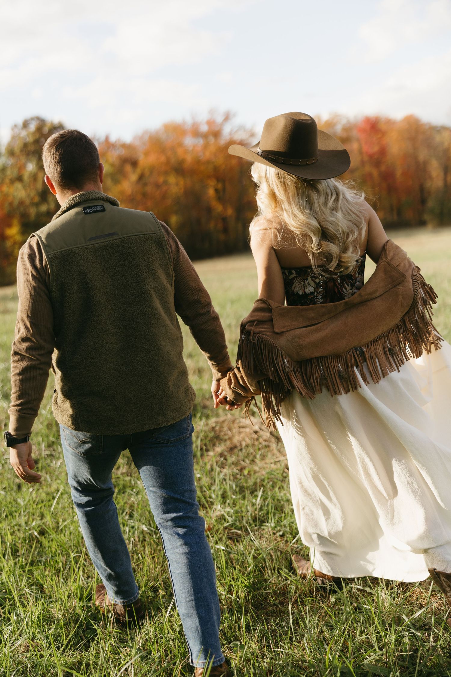A couple walks hand-in-hand through a green field, the woman wearing a fringed jacket, cowboy hat, and white dress.