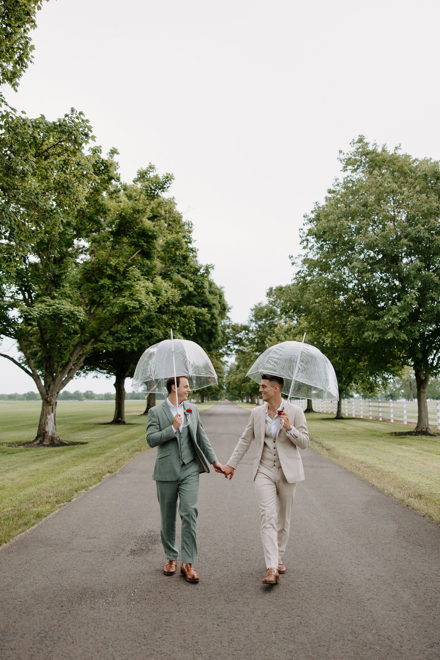 Two brides in suits hold clear umbrellas while walking hand in hand down a tree-lined path on their wedding day.