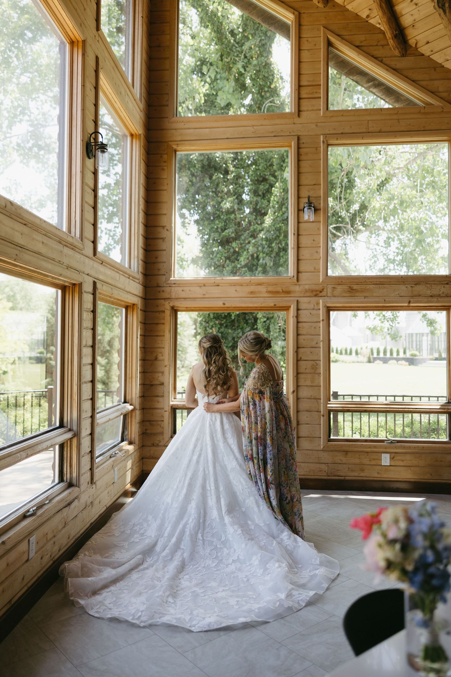 Bride in white gown with bridesmaid in floral dress standing by large wooden cabin windows.