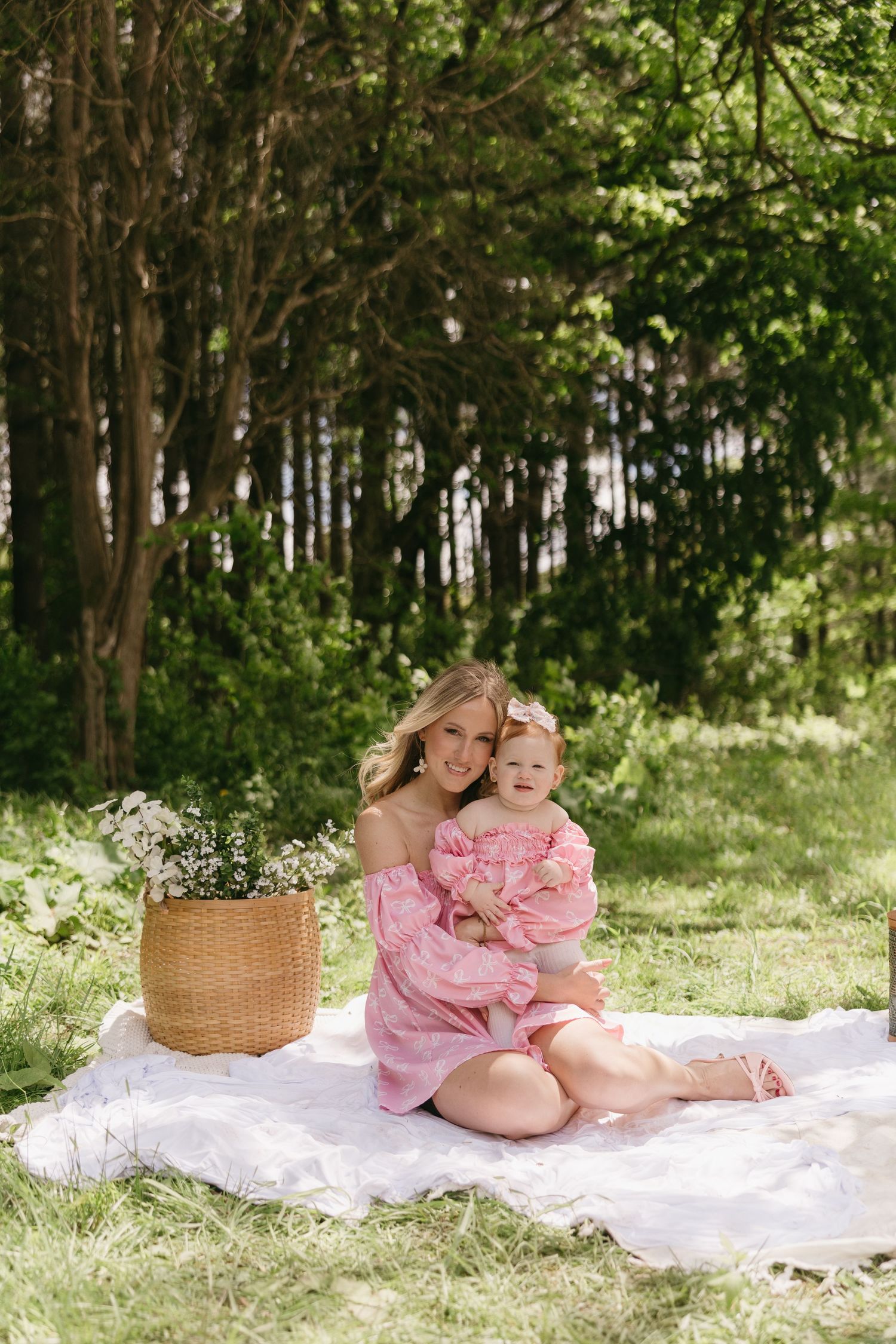 Mother and baby girl in matching pink dresses sitting on white blanket in green park with wicker basket of flowers.
