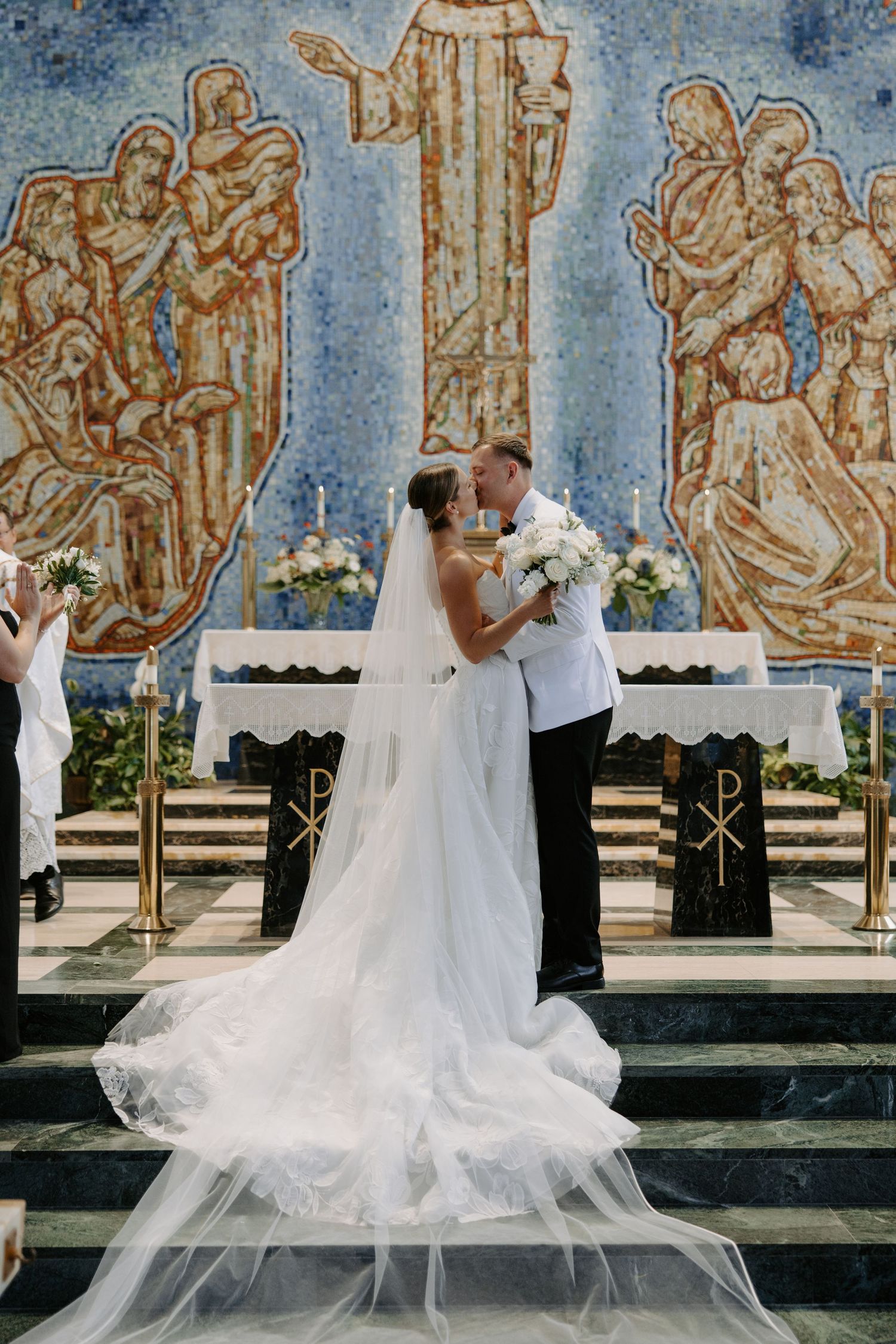 Bride and groom share a kiss at the altar of a church adorned with a stunning mosaic mural backdrop.