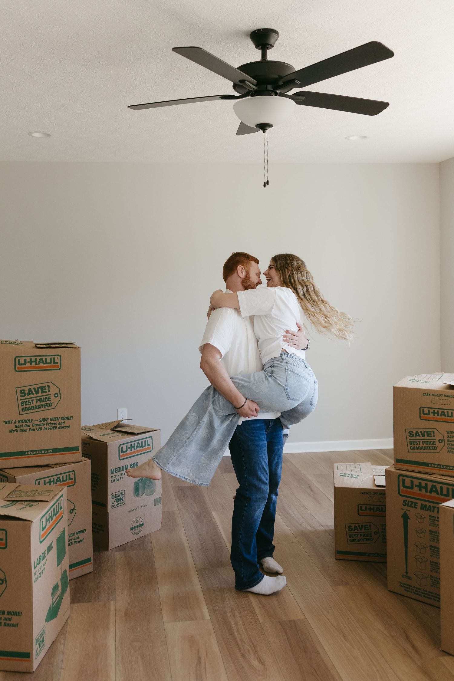 Couple sharing a joyful lift and kiss surrounded by moving boxes in their new empty home.