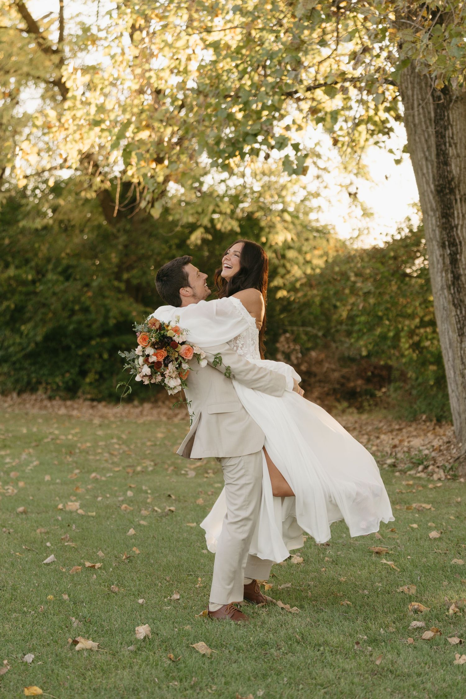 Joyful groom lifting laughing bride in white dress outdoors among autumn trees.
