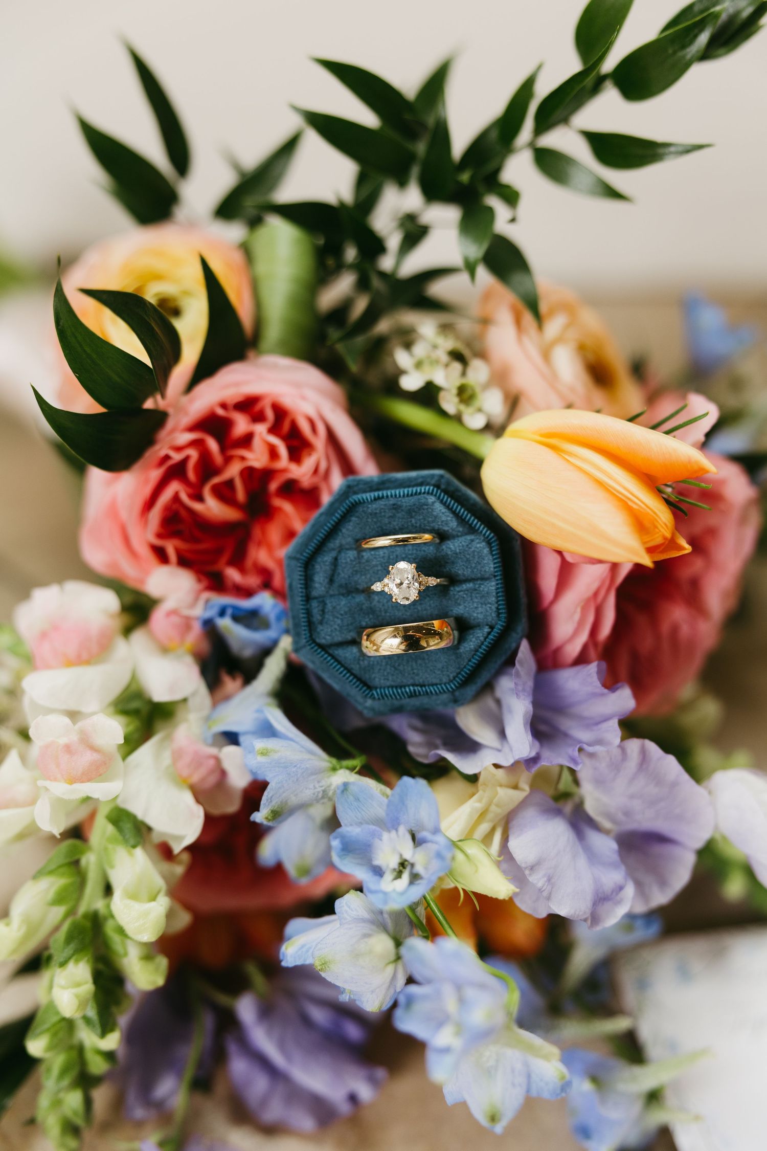 Wedding rings displayed in a teal velvet box surrounded by a vibrant colorful bridal bouquet.