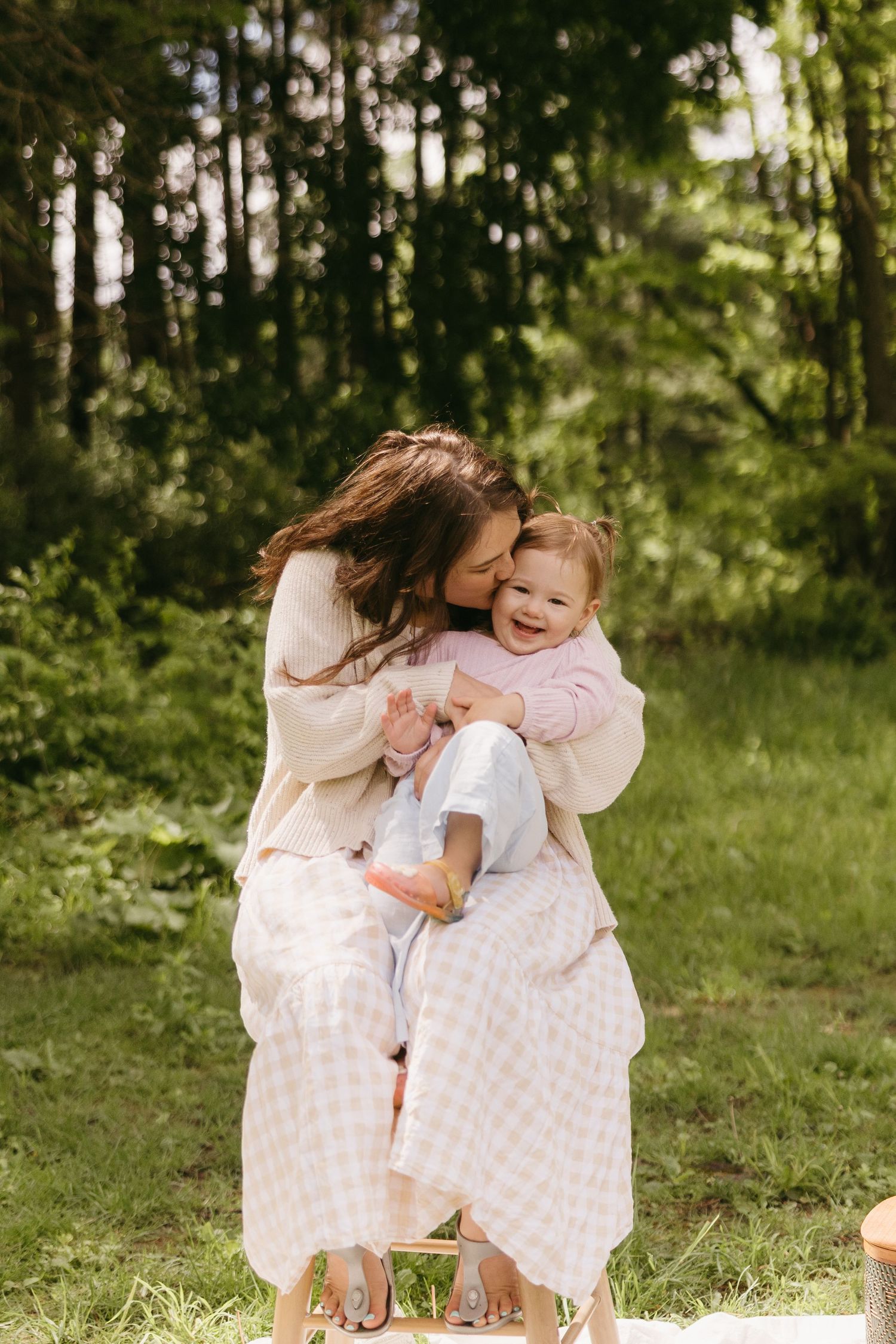 A mother kisses her laughing toddler outdoors in a lush green forest setting, both wearing soft pastel outfits.