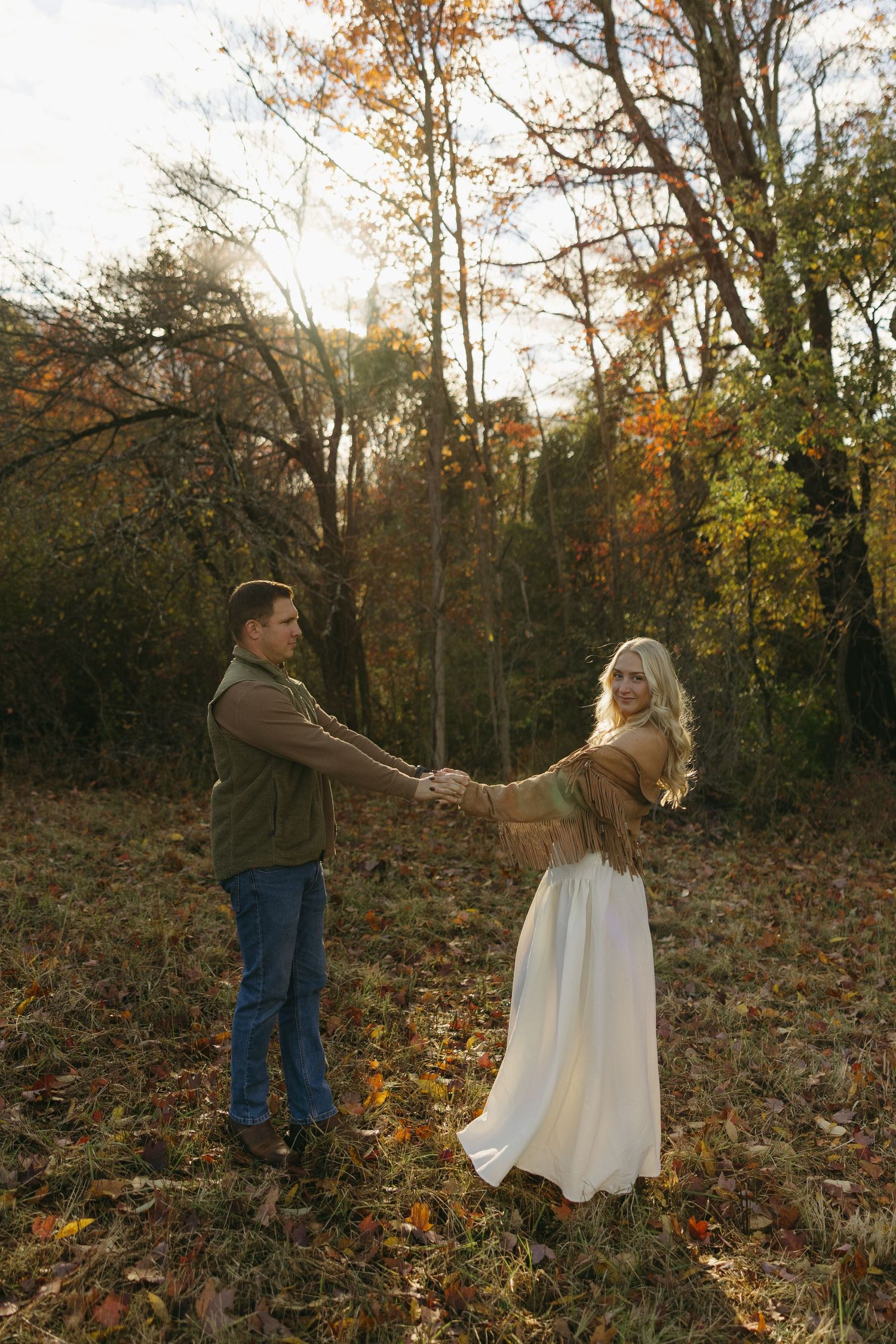 Couple holding hands in an autumn forest clearing, woman in white skirt and brown shawl, man in green jacket and jeans.