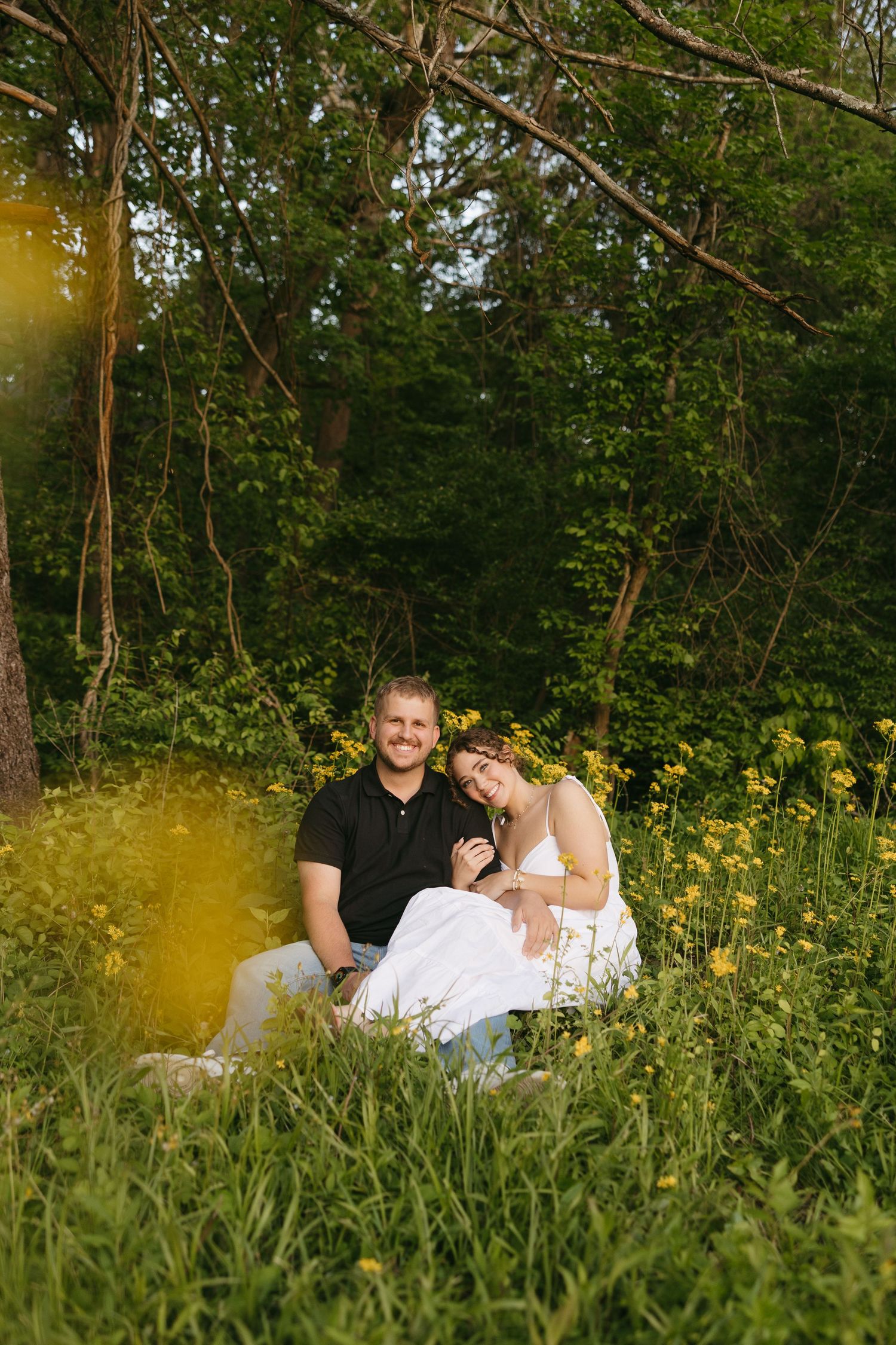 A smiling couple sits together in a lush green field of yellow wildflowers, surrounded by trees in warm golden sunlight.