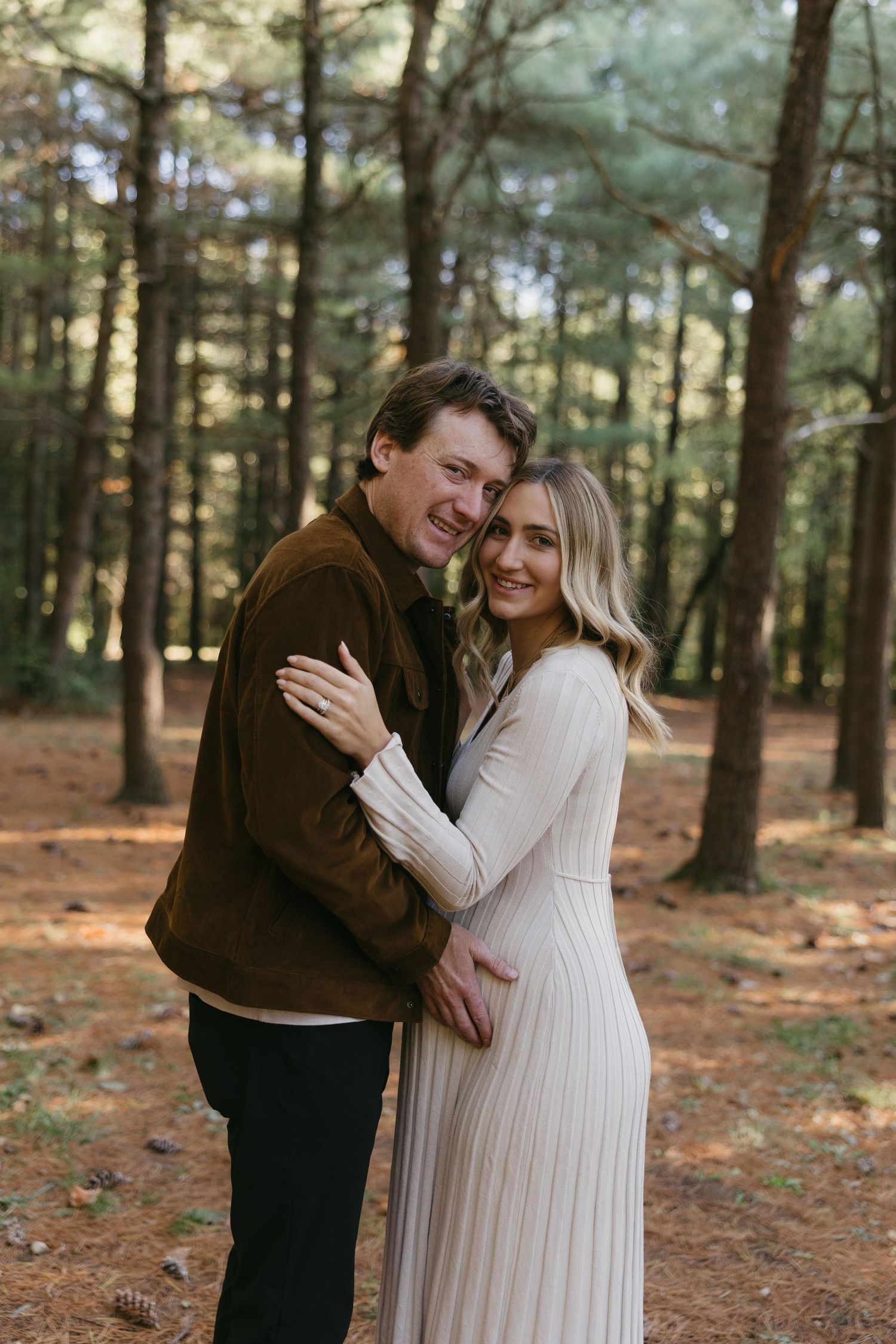 Couple embracing in a pine forest, woman in white ribbed dress and man in brown jacket, smiling at camera.