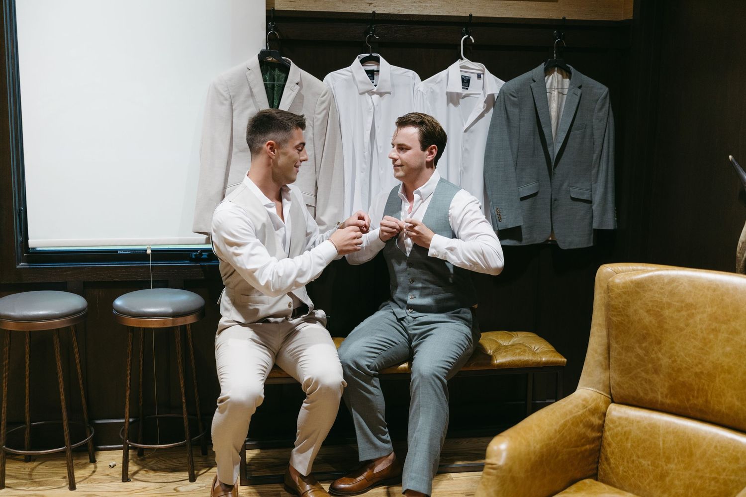 Two well-dressed men sit together in a stylish showroom, exchanging items near hanging suits and shirts.