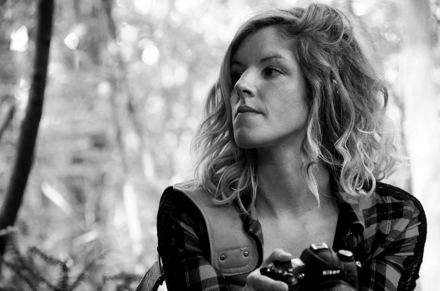 Black and white portrait of a curly-haired woman holding a Nikon camera outdoors in nature.