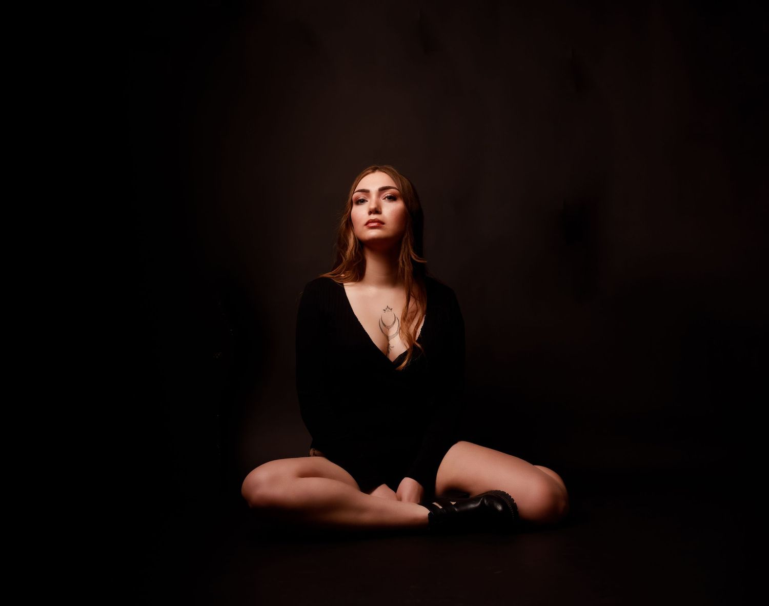 Young woman in black deep-V bodysuit sits cross-legged on floor in dramatic dark studio setting.