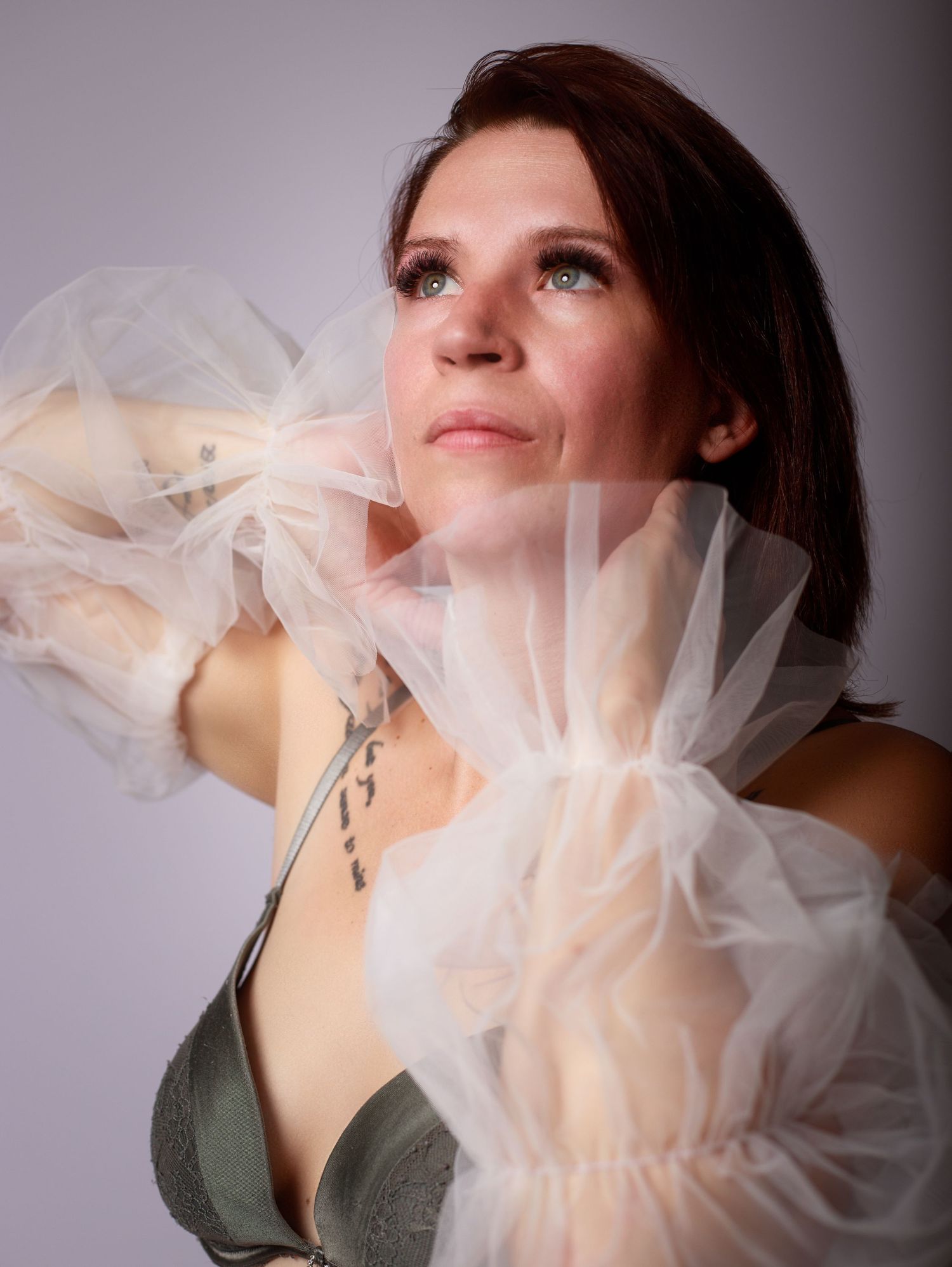 Woman in sage green bra holds sheer white tulle fabric looking upward in soft studio light.