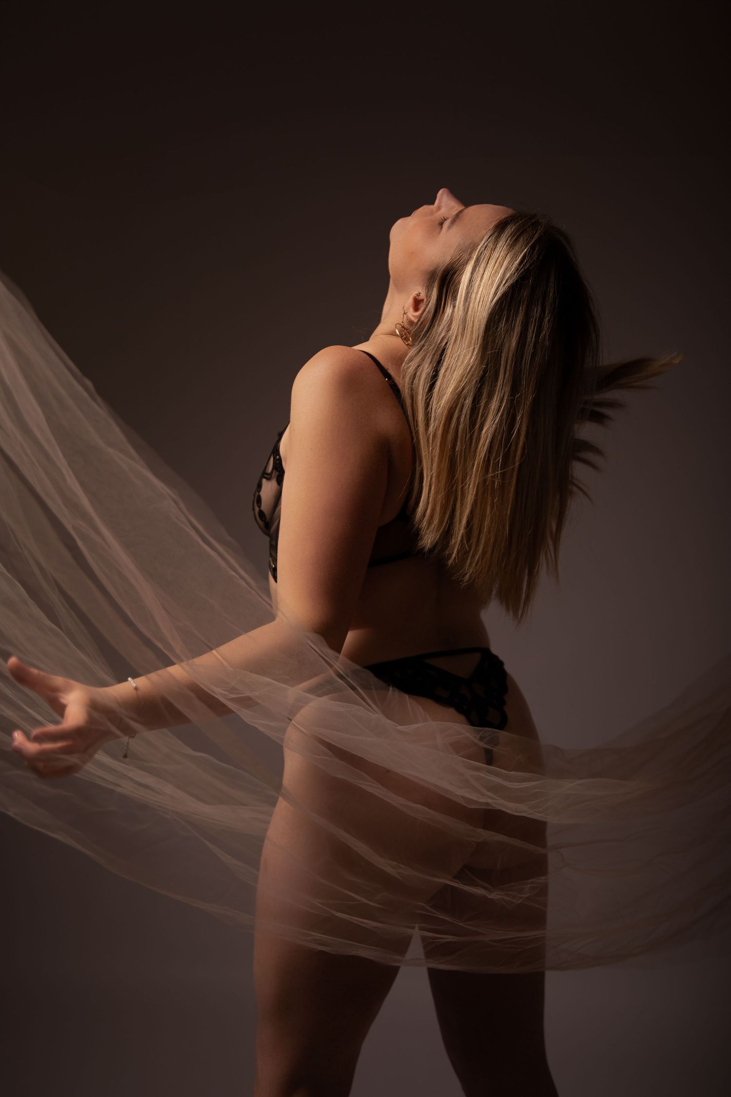 Blonde woman in black lingerie dancing with flowing sheer fabric on dark studio background.