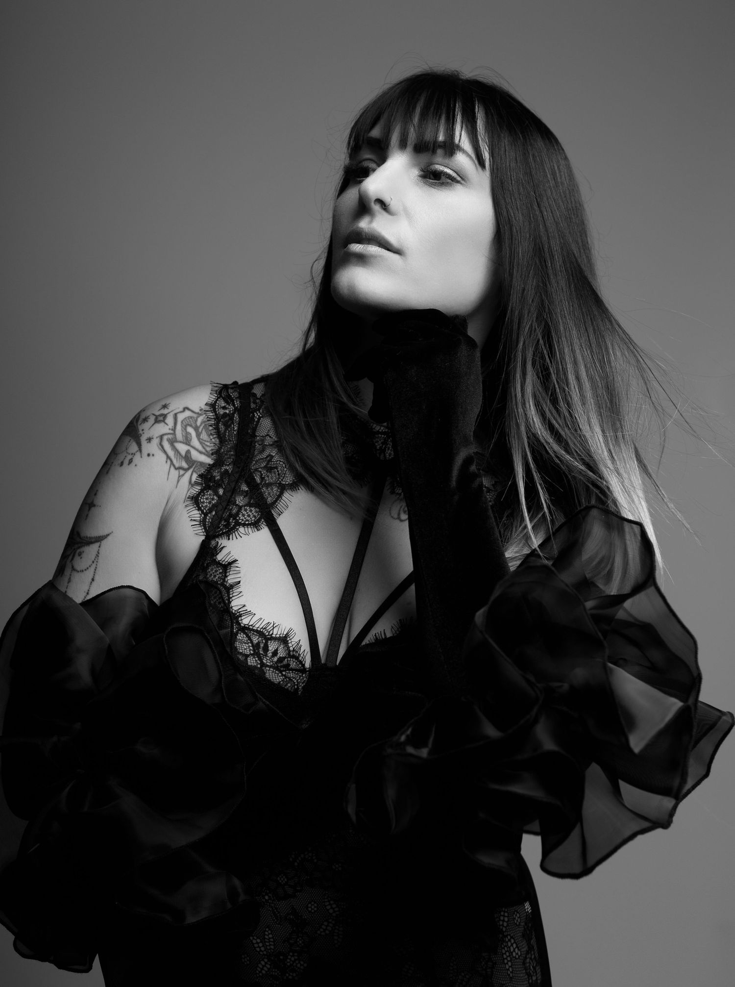 Black and white portrait of dark-haired woman in black lace bodysuit with ruffled sleeves and arm tattoo.
