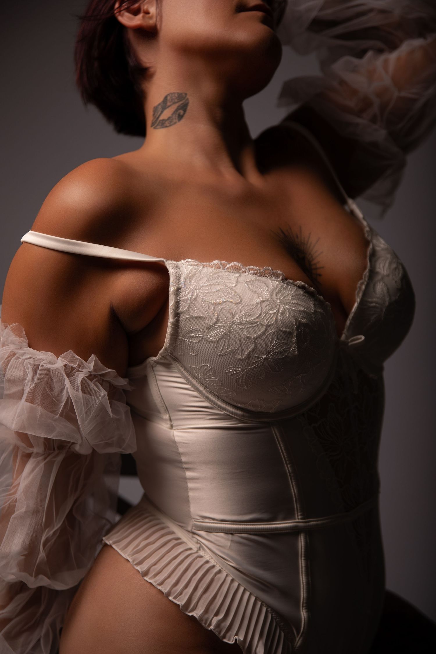 Woman in white lace corset with sheer ruffled sleeves and a lip tattoo on her neck, studio portrait.