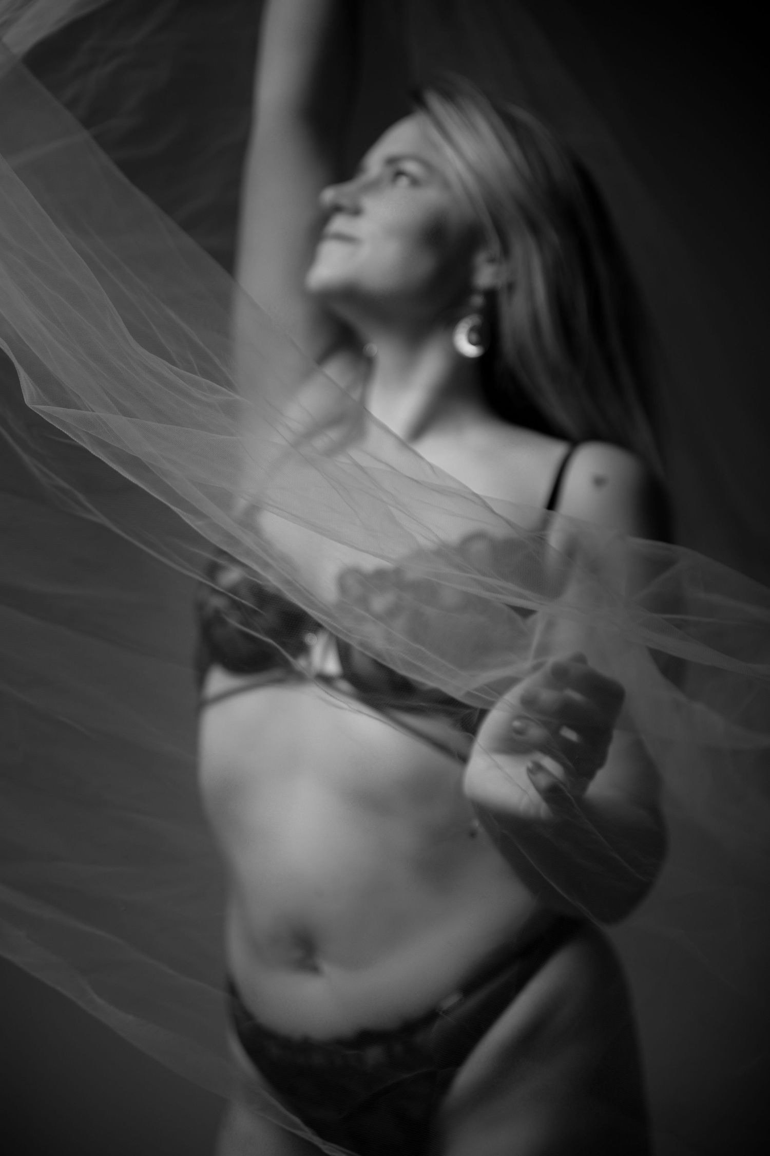 Black and white boudoir photo of woman in lingerie holding sheer fabric with arm raised.