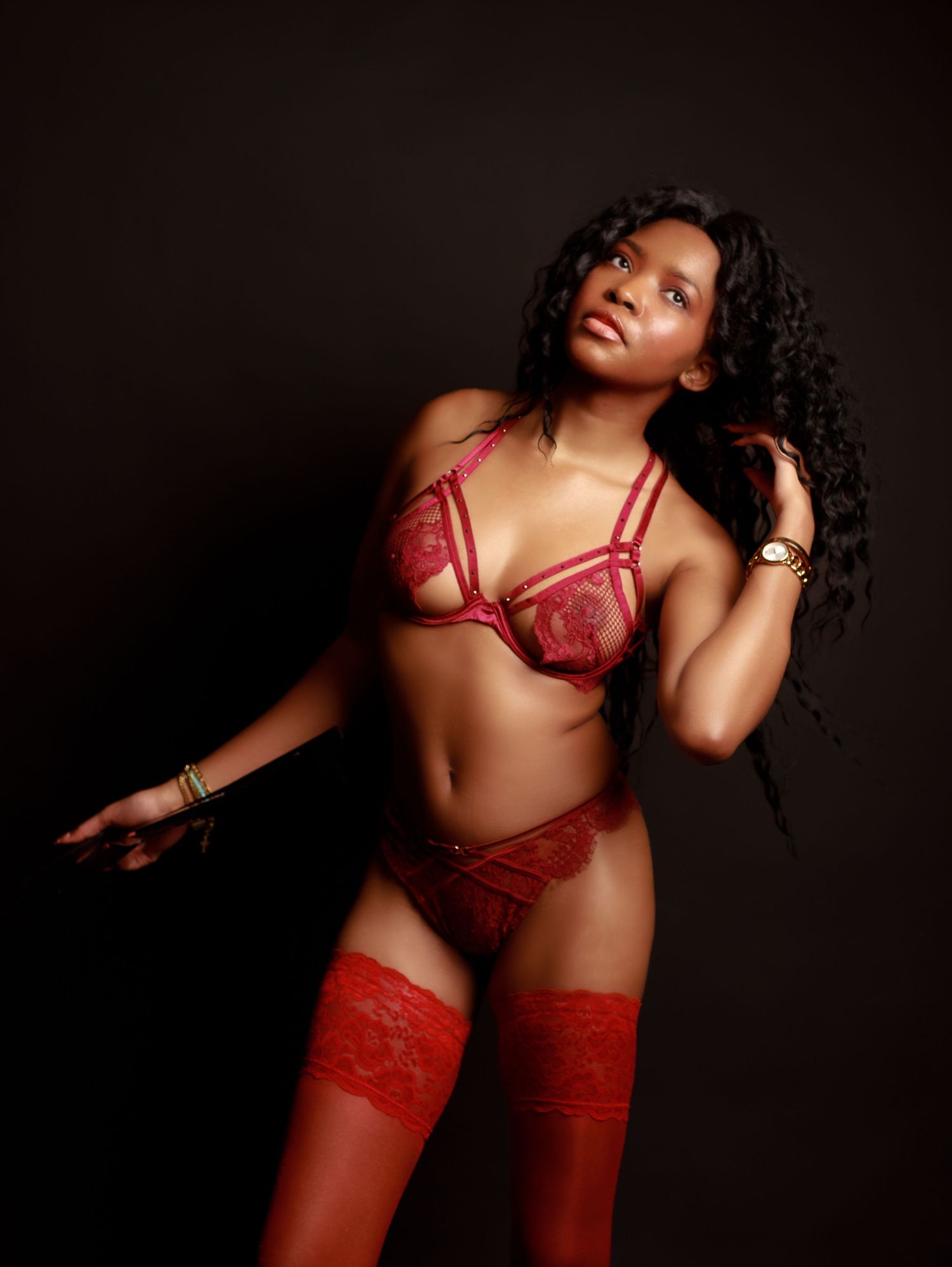 Woman in red lace lingerie set with matching thigh-high stockings poses against dark background.