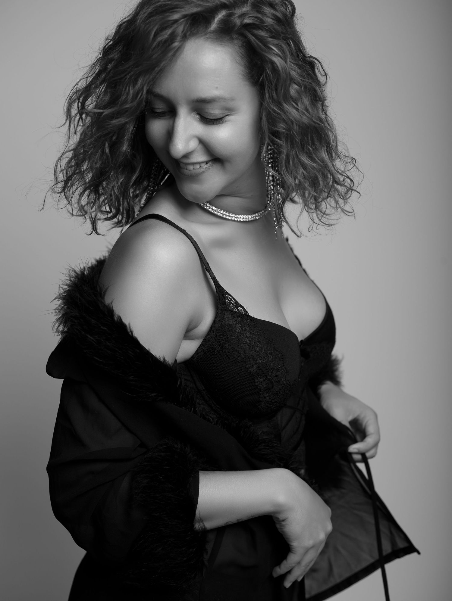 Black and white portrait of a smiling woman in black lace lingerie and fur-trimmed robe.