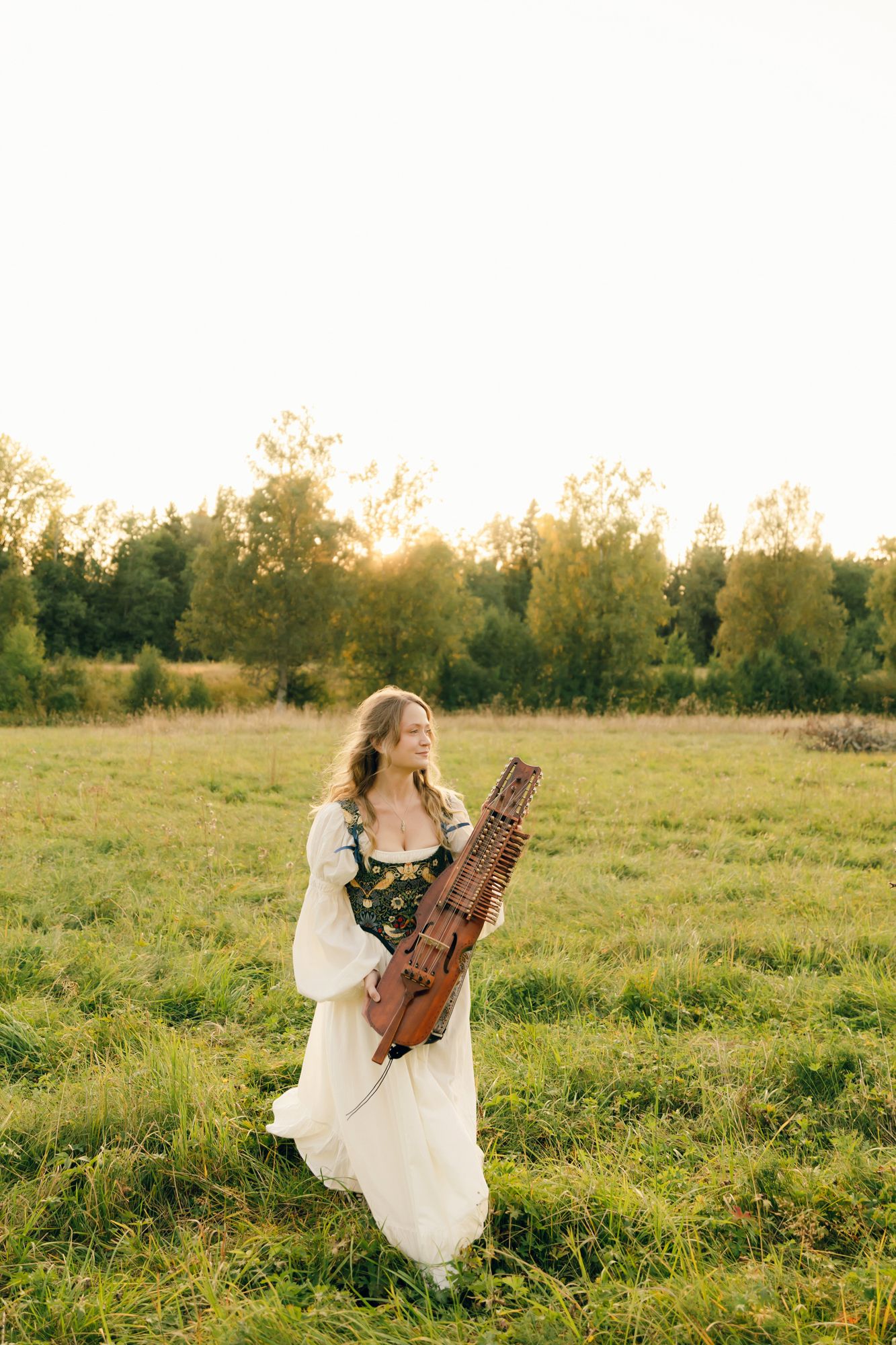 Porträtt på Julia Johansson i Karlstad. Julia går på ängen med sin nyckelharpa i vit klänning.