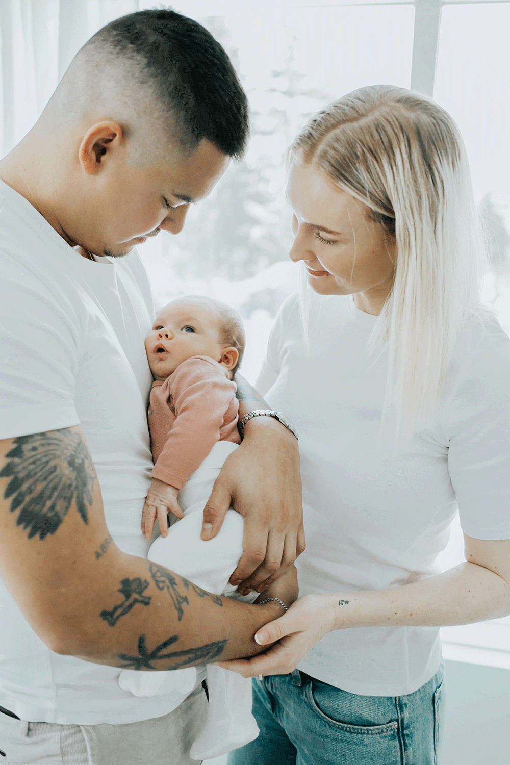 Familjefotografering. Två föräldrar och deras bebis. Föräldrarna är klädda i vitt och bebis i rosa.