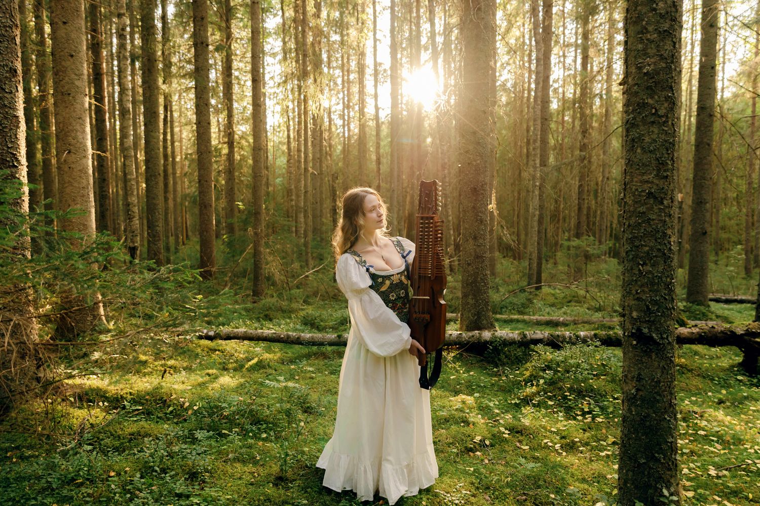 Porträtt på Julia Johansson i Karlstad. Hon står i skogen tillsammans med sin nyckelharpa. Solen i bakgrunden.