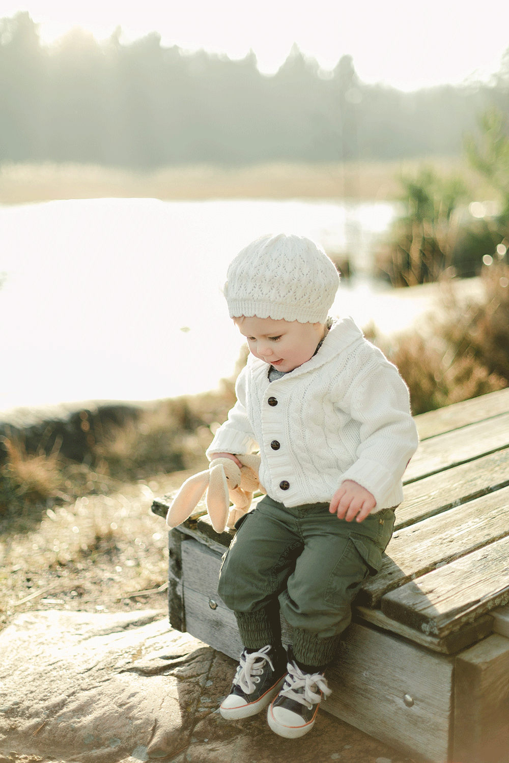 Bebis som sitter med sitt gosedjur på en brygga framför en sjö på vintern. Bebisen har mössa och Converse.
