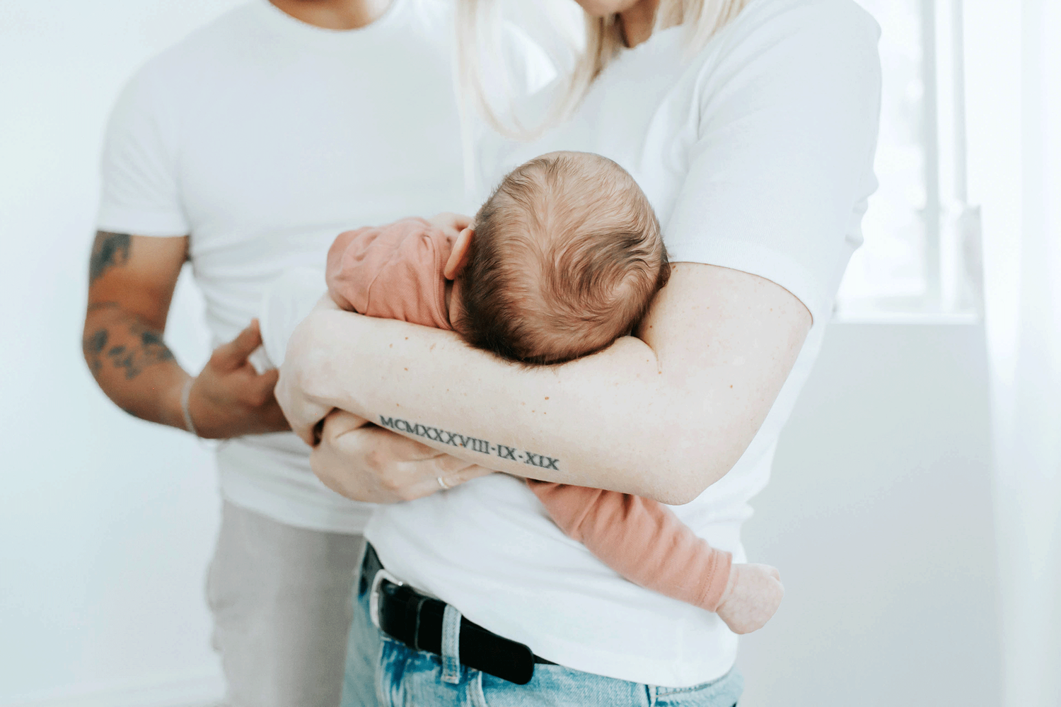 Ljus bild på bebis i mammas famn. Ser bara bakhuvudet på bebisen. Pappan står i bakgrunden.