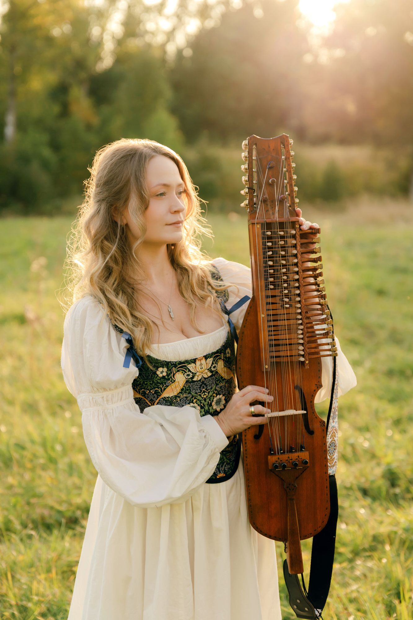 Porträtt på Julia Johansson i Karlstad. Julia står på äng tillsammans med sin nyckelharpa som hon håller bredvid sig. Halvkroppsbild.