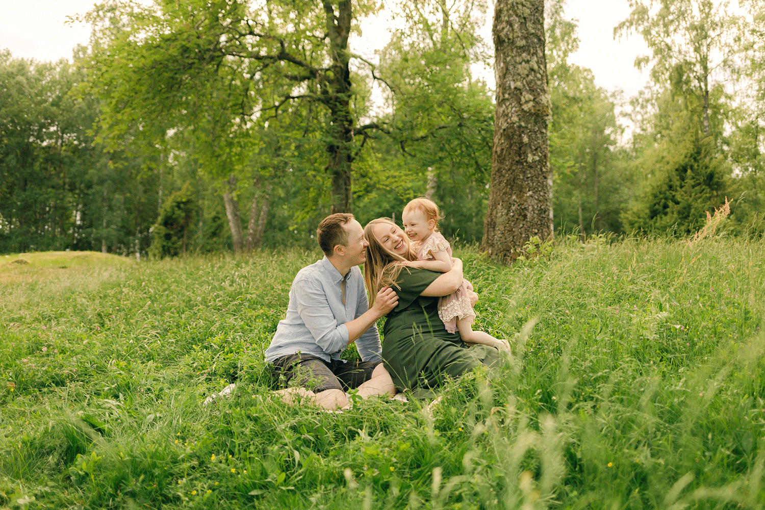 Familjefotorgafering. Sitter på en grönskande äng. Dottern i mammas famn och alla skrattar.