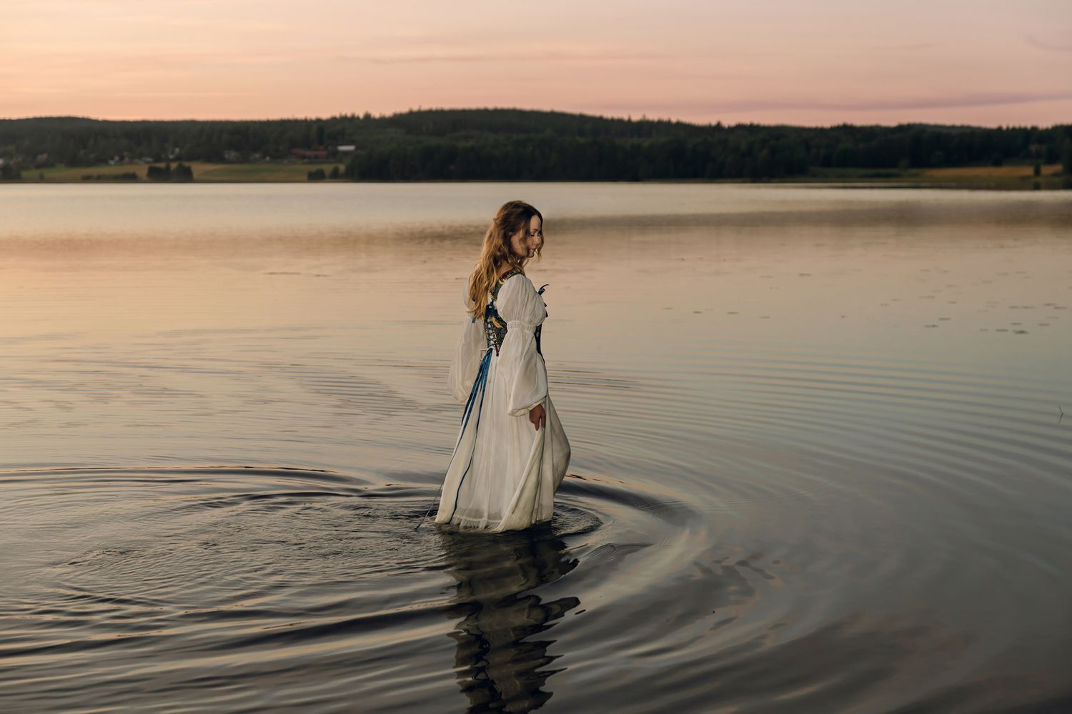 Porträtt på Julia Johansson i Karlstad. Stående i spegelblank sjö. Rosa och lila himmel. Hon går i vattnet.