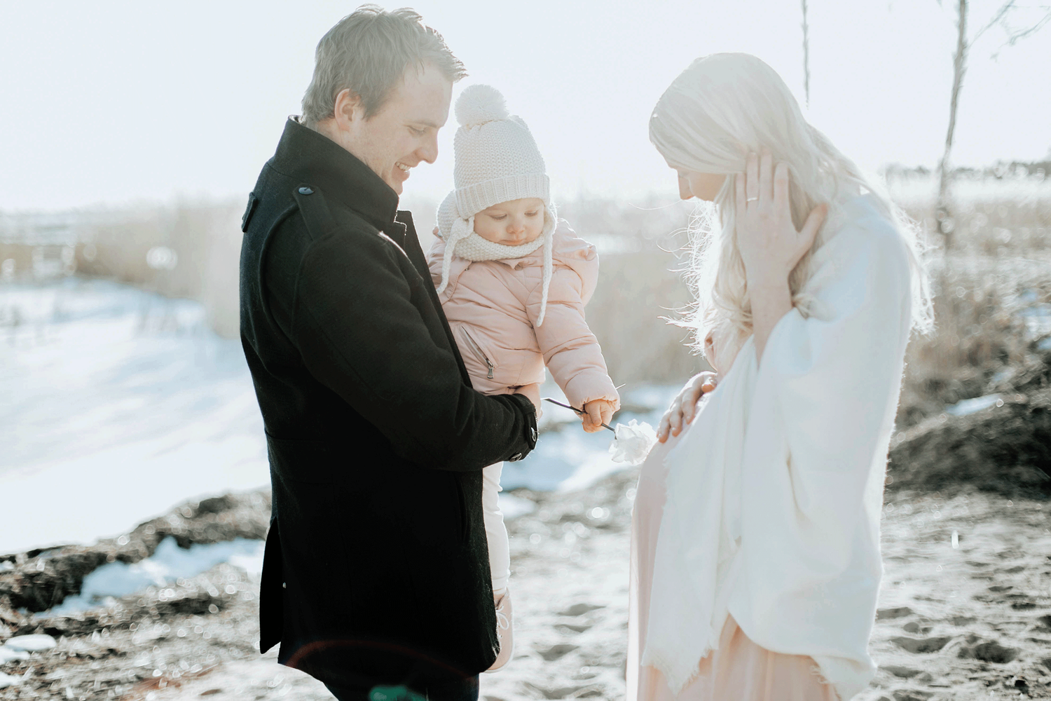 Familjefotografering i Alster. Mamman gravid och pappan håller i dottern. Dottern håller i en blomma som hon pekar på mammas mage med.