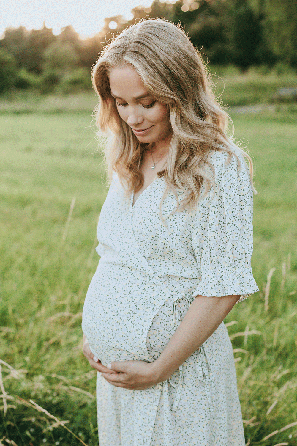 Gravid kvinna i solnedgång på en äng. Hon tittar ner mot magen och håller under den. Ljus klänning på sig.