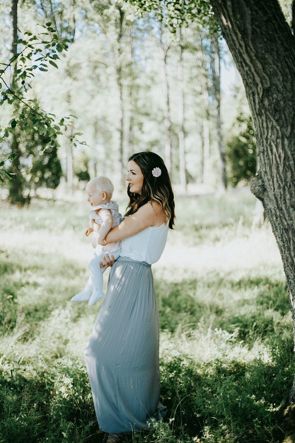 Mamma som bär sitt barn under ett träd. Mamman har kjol och blomma i håret. Barnet skrattar.