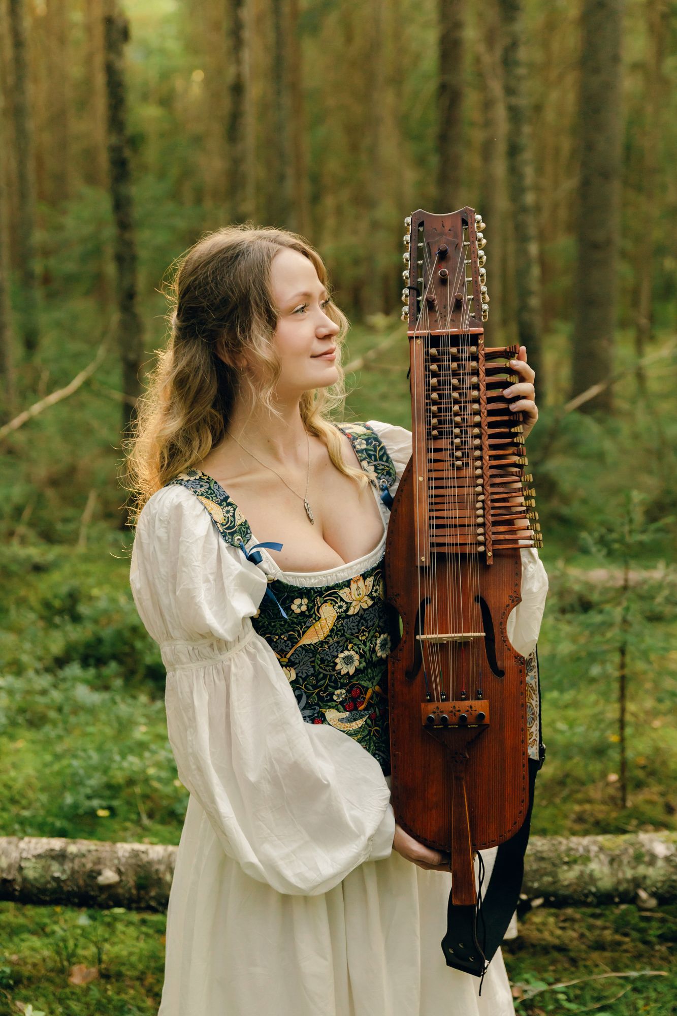 Porträtt på Julia Johansson i Karlstad. Hon står med sin nyckelharpa bredvid sig i skogen.