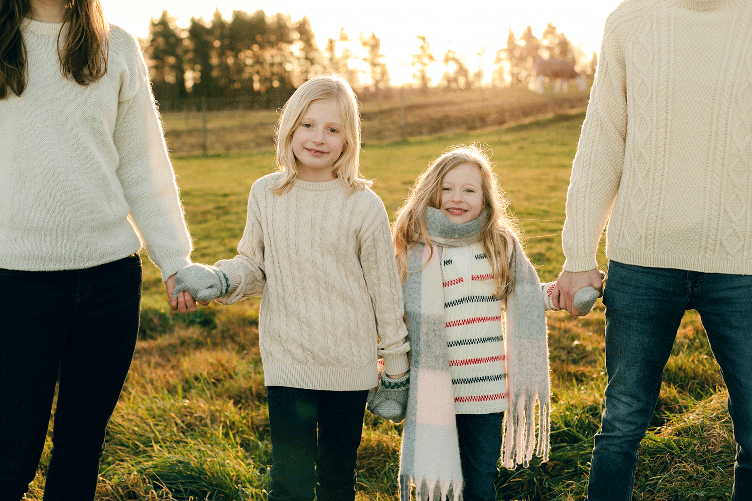 Familjefotografering i solnedgång på en åker. Bara barnen syns men de håller i föräldrarnas händer. Alla har ljusa färger upptill.