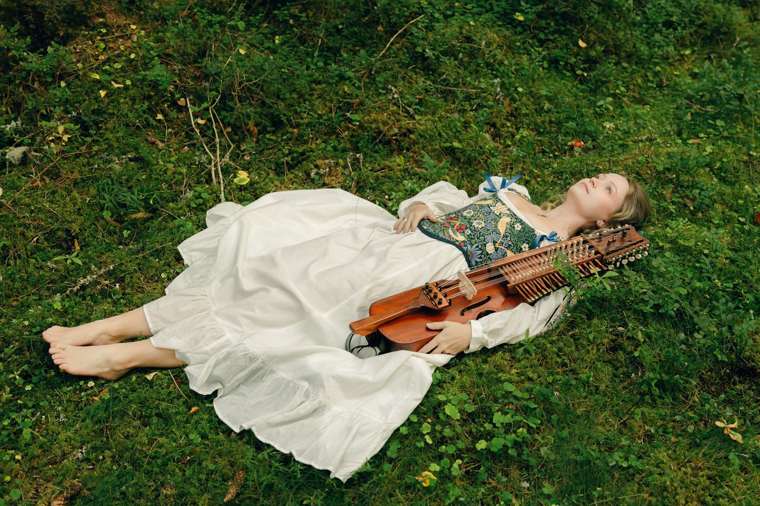 Porträtt på Julia Johansson i Karlstad. Ligger bland mossan i skogen med sin nyckelharpa bredvid sig. Helkroppsbild.