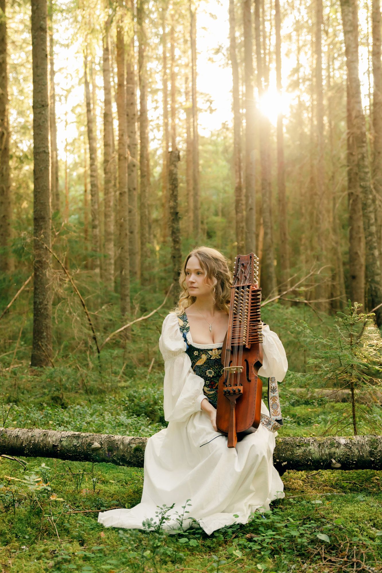 Porträtt på Julia Johansson i Karlstad. Hon sitter med sin nyckelharpa i knät i skogen på ett fallet träd.