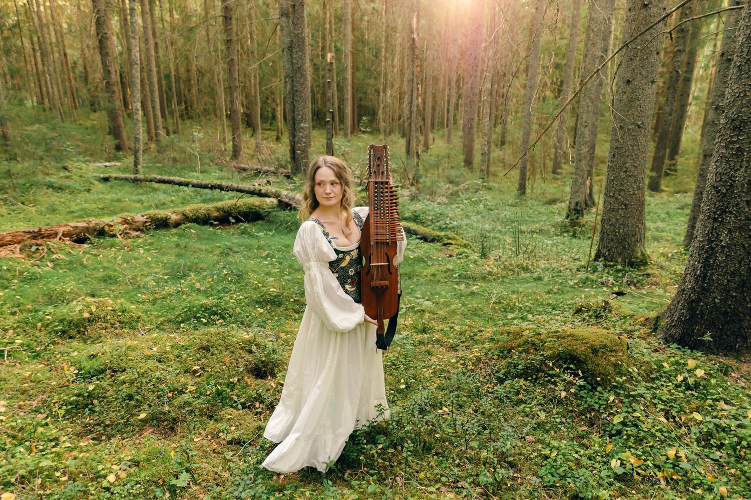 Porträtt på Julia Johansson i Karlstad. Hon håller sin nyckelharpa och står i skogen i vit klänning.