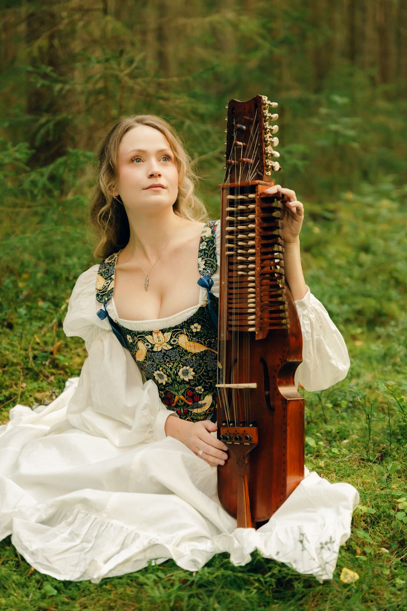 Porträtt på Julia Johansson i Karlstad. . Hon sitter i skogen bland mossan med sin nyckelharpa bredvid sig. Tittar upp mot träden.