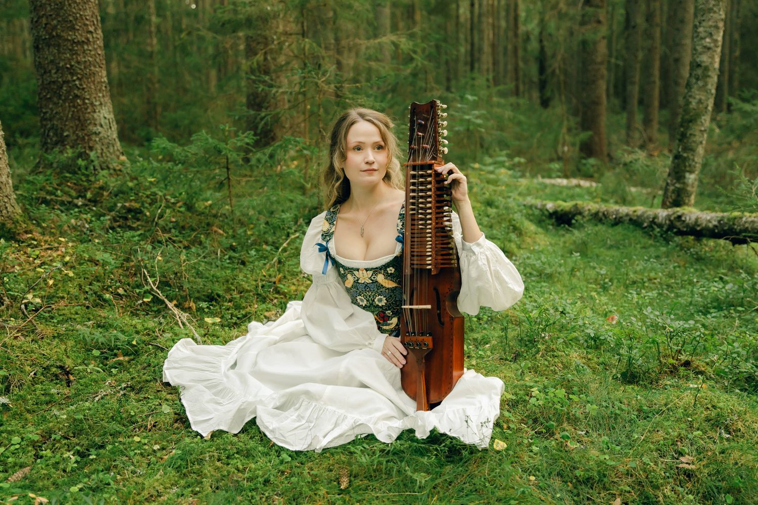 Porträtt på Julia Johansson i Karlstad. Hon sitter i skogen bland mossan med sin nyckelharpa bredvid sig.
