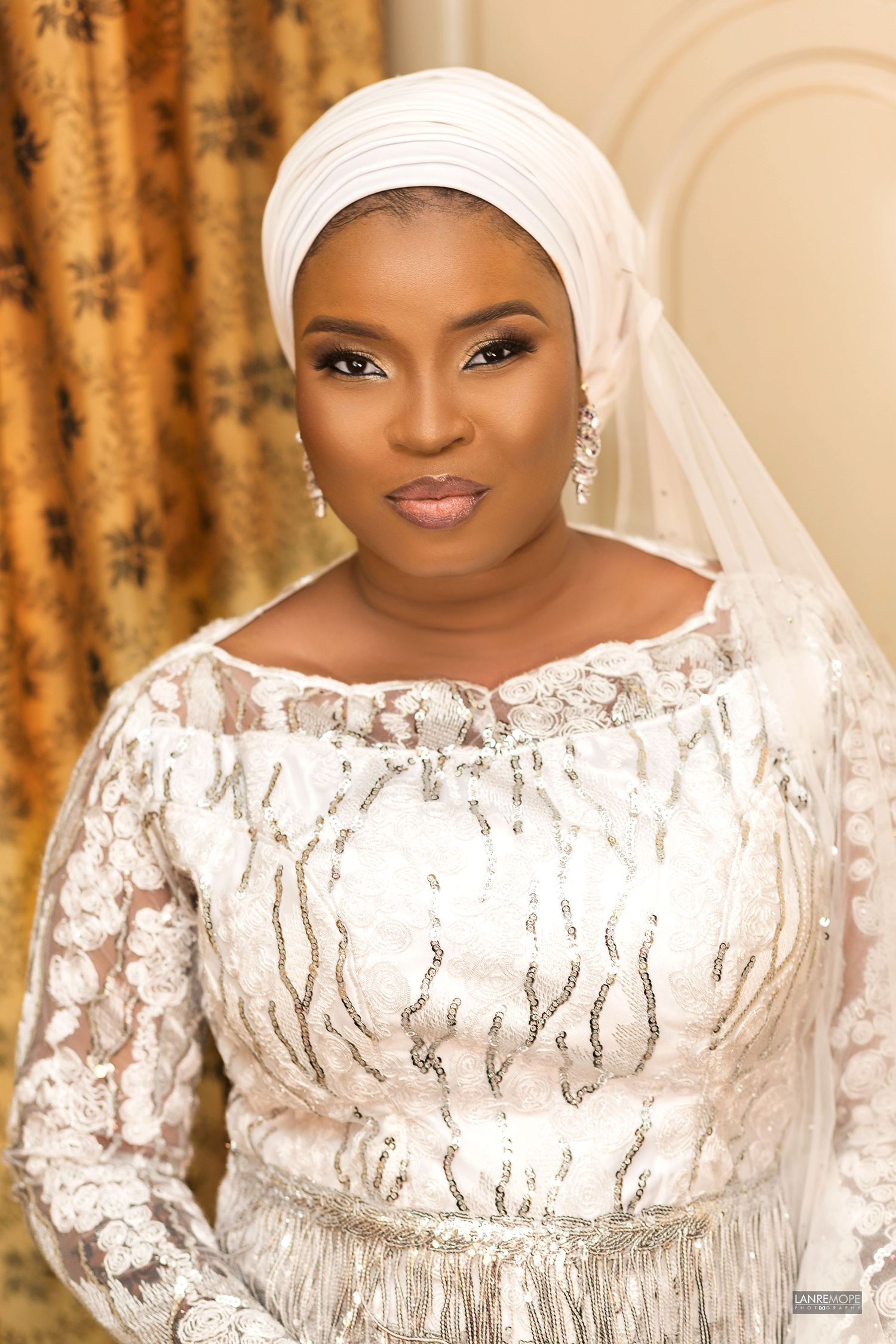 Elegant bride in white lace gown and hijab with veil, wearing crystal earrings, posing for wedding portrait.