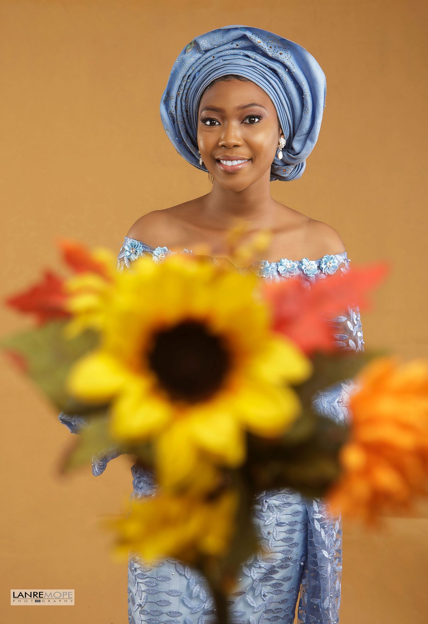 Woman in blue lace off-shoulder dress and gele headwrap holding sunflowers in studio portrait.