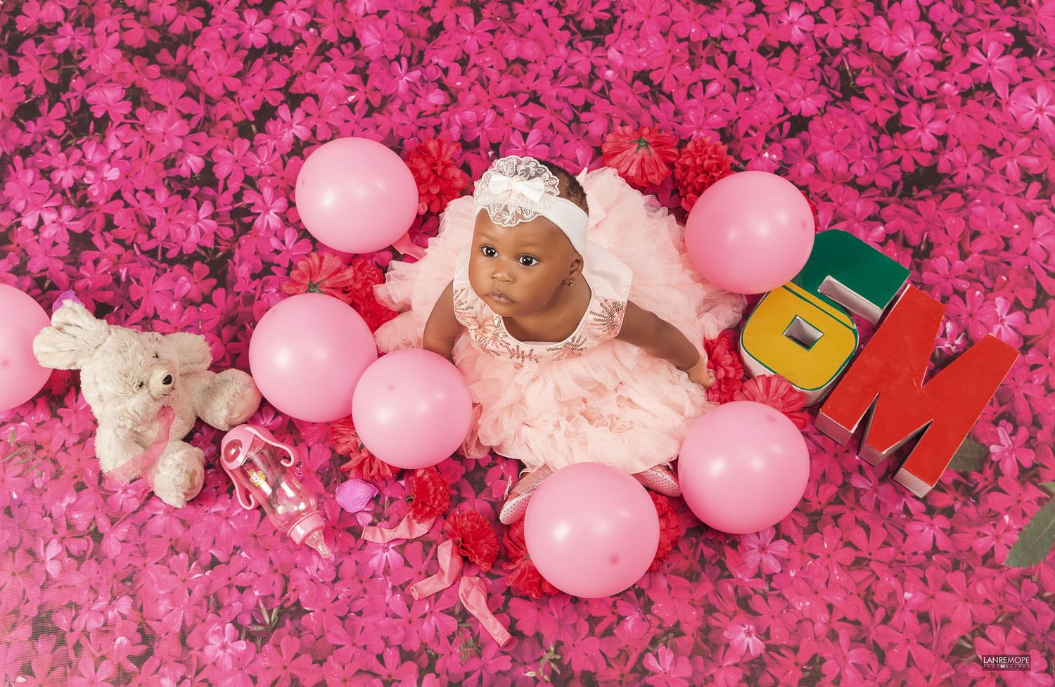 Newborn baby in pink lace outfit surrounded by pink balloons and flowers in a milestone photo shoot.