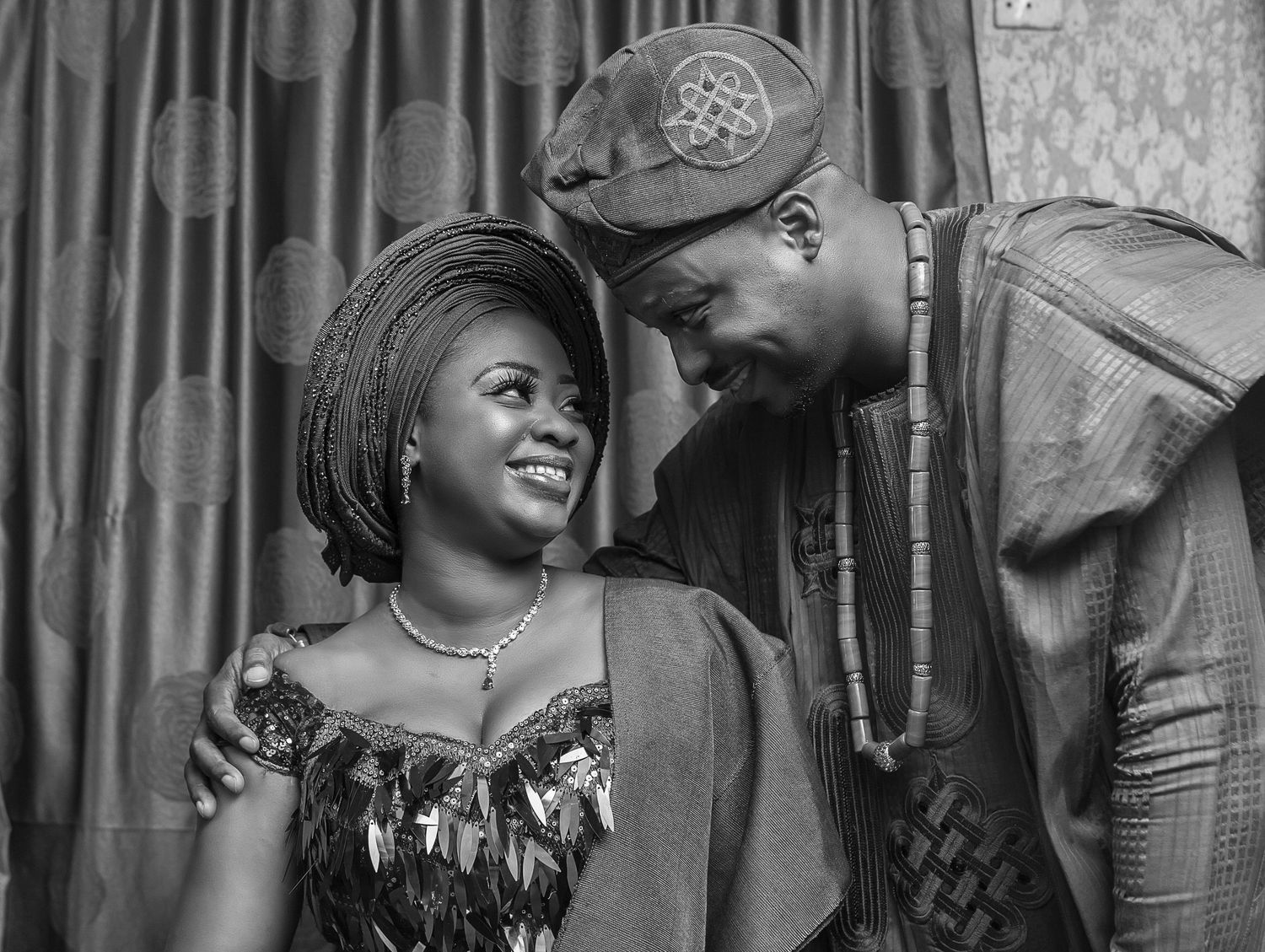Nigerian couple in traditional attire smiling at each other in black and white photo.