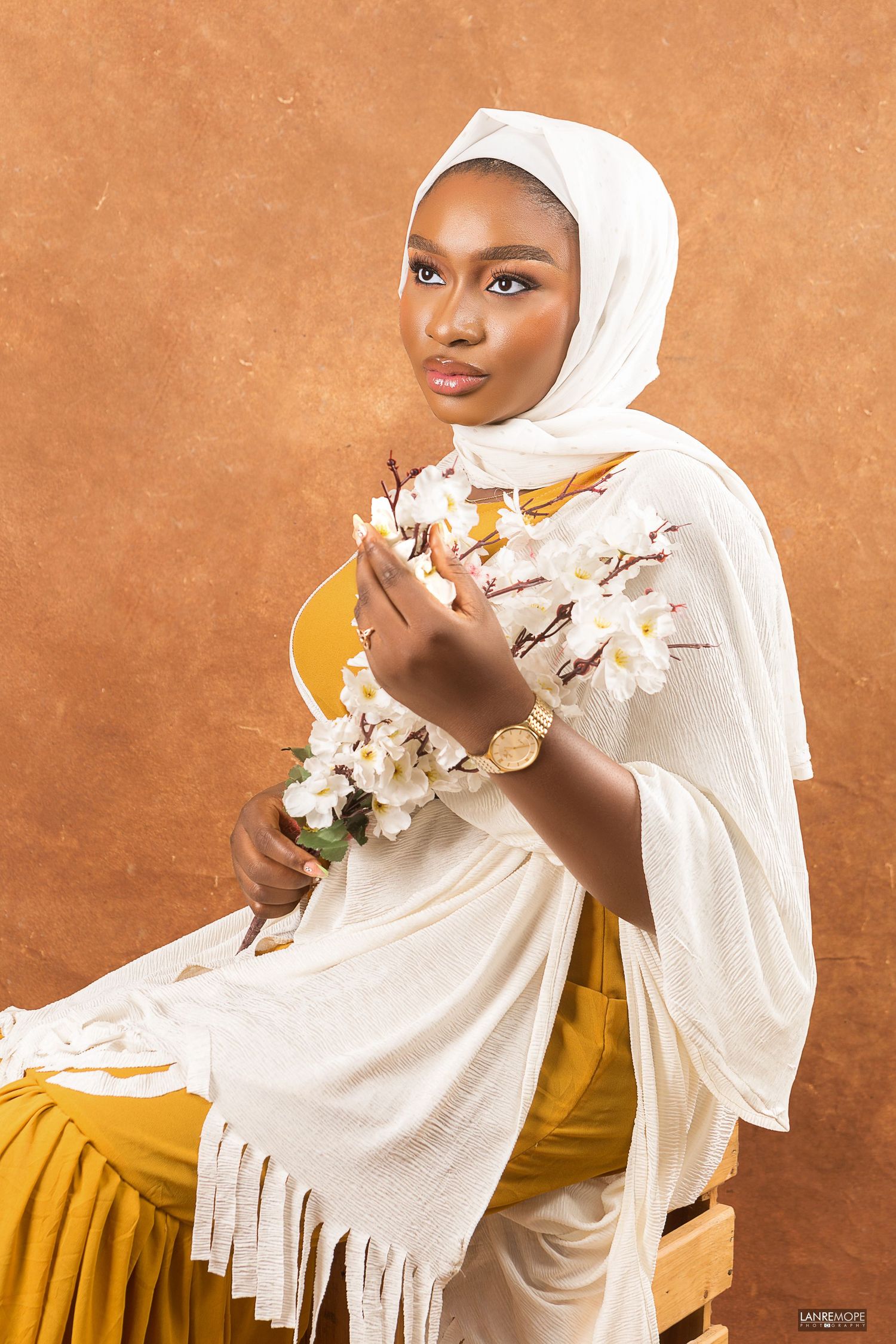 An elegant woman in a white hijab and outfit holds white flowers while posing for a studio portrait.