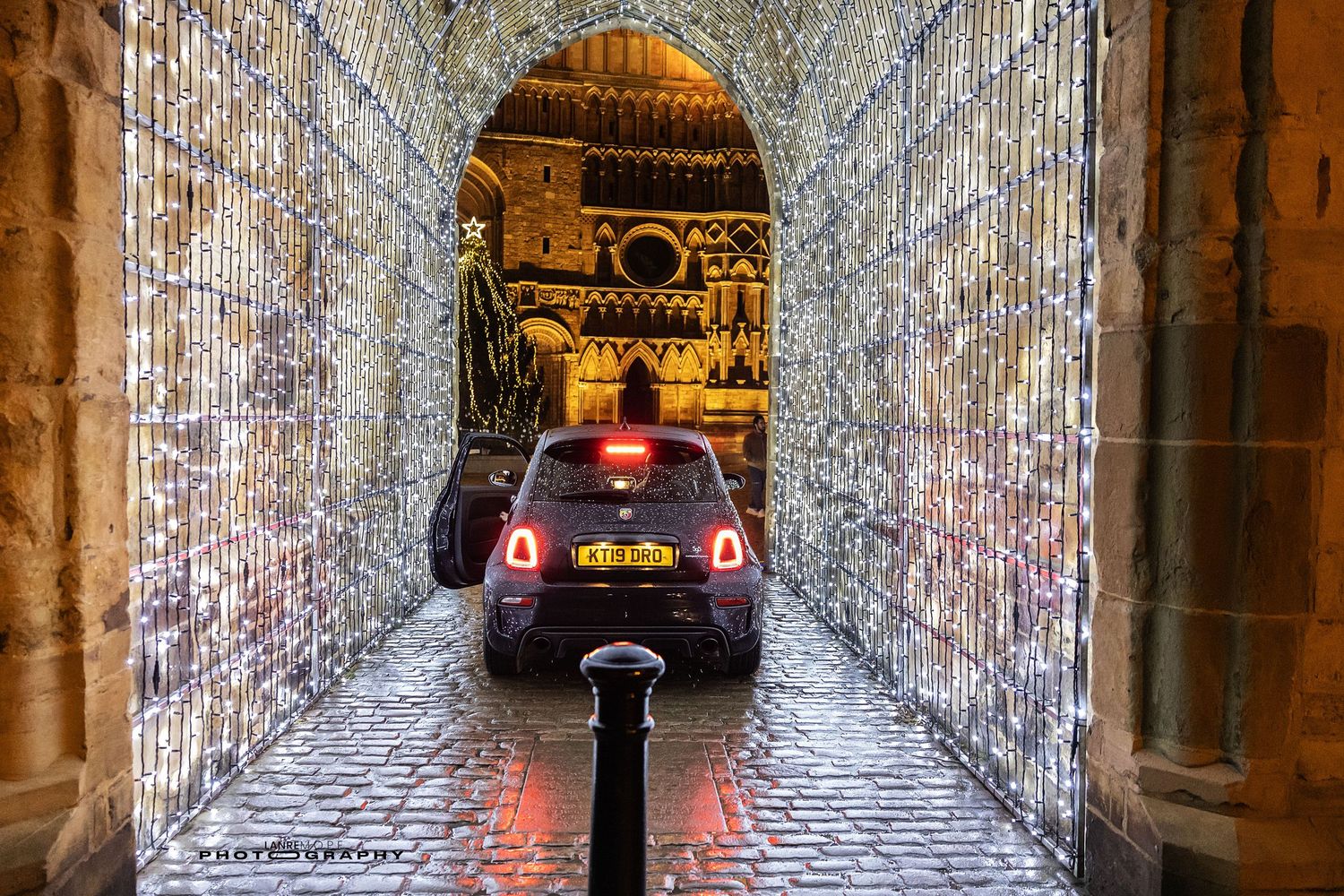 Car driving through a festive illuminated archway tunnel with Christmas lights at night in a historic stone building.
