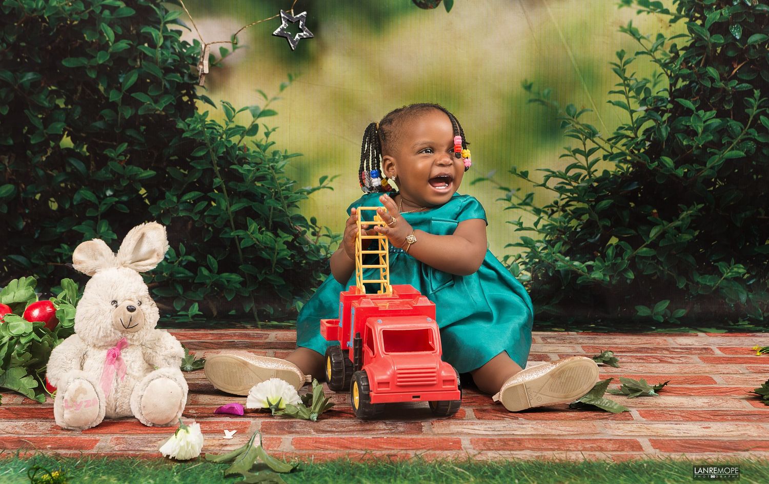 Happy toddler girl in teal dress playing with red toy truck outdoors in a garden photo session.